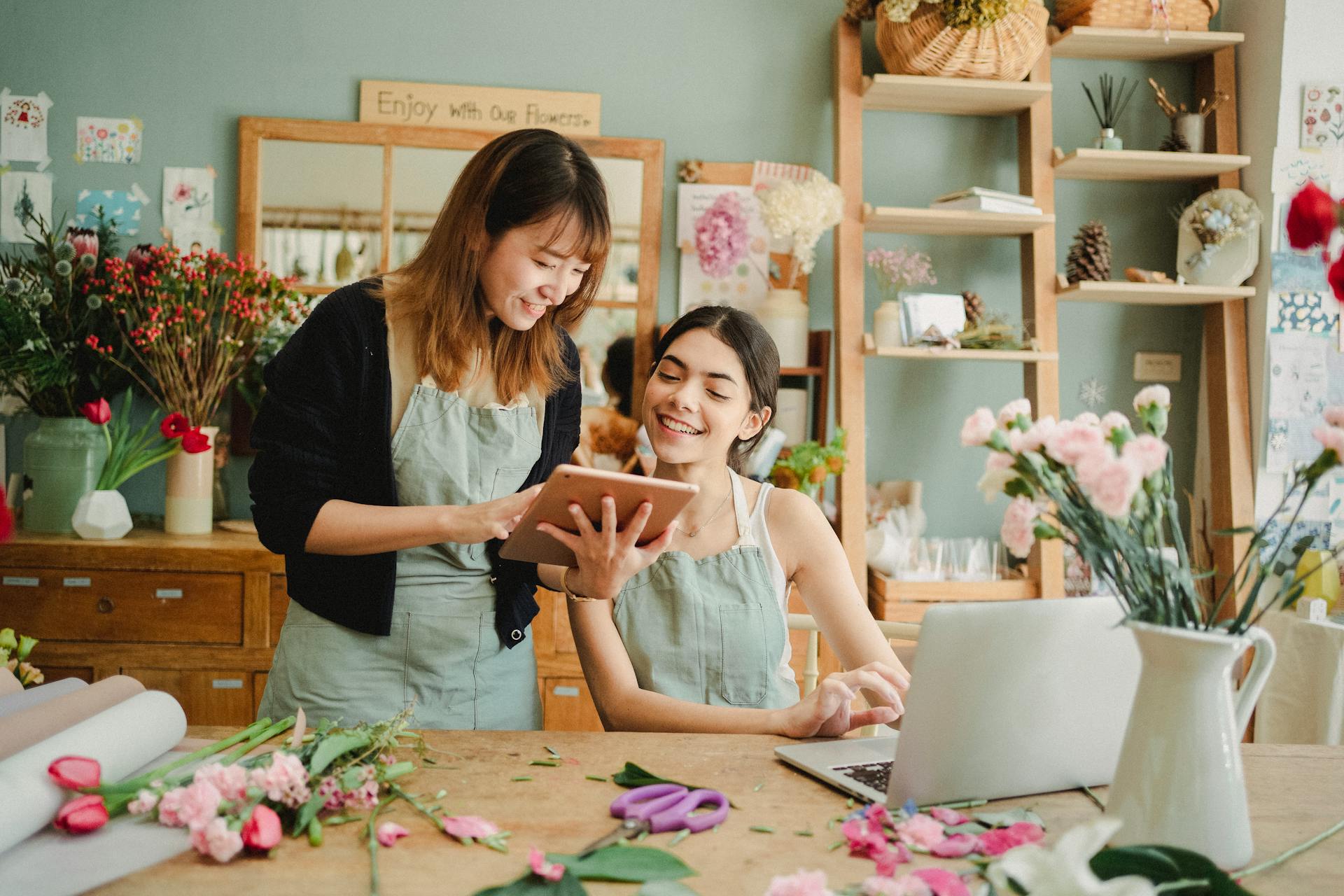 Content multiethnic female florists in aprons at table with laptop and browsing tablet while preparing for order in creative floristry studio