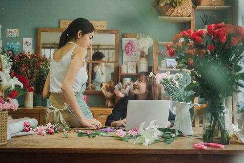 Free Content multiethnic female colleagues sitting at table with bouquets of flowers and working on computer in florist shop while looking at each other Stock Photo