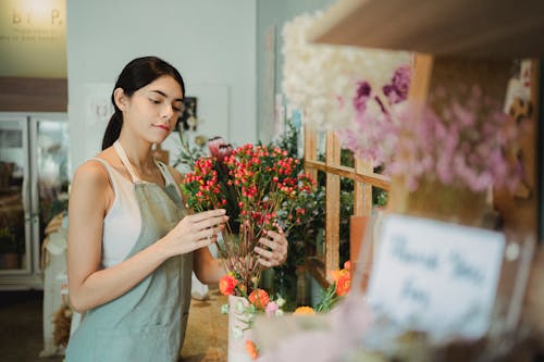 Positive female florist with dark hair making composition of small red flowers while preparing order during work in floristry studio