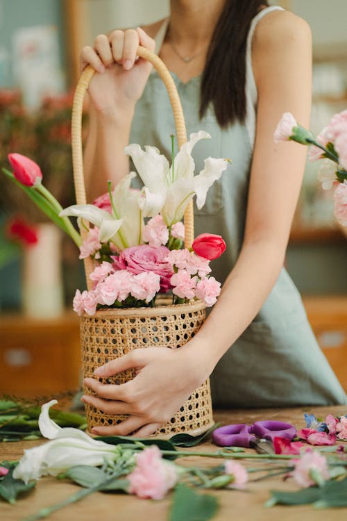 Unrecognizable female florist in apron standing at table with bouquet of flowers in wicker basket in floral store during work