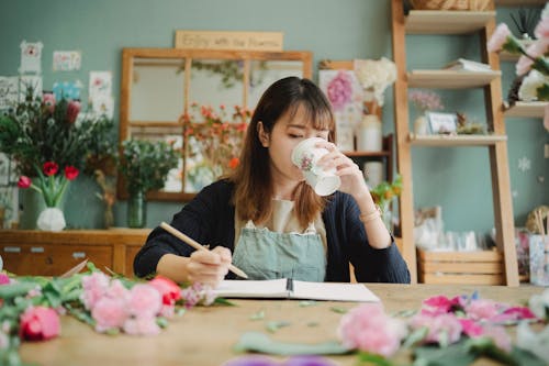 Concentrated Asian female florist drinking hot beverage and sitting at table with flowers while taking notes in notebook during work
