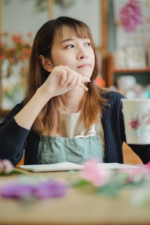 Pensive Asian florist looking away while sitting at table with hand near face and cup of hot beverage in floristry store