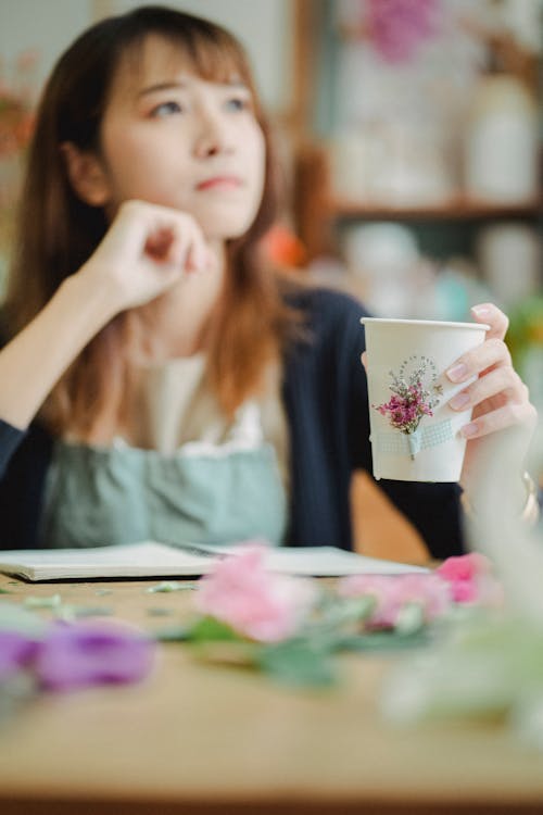 Wistful Asian female florist with hand near face looking away while sitting at table with flowers and notebook with cup of aromatic beverage in hand