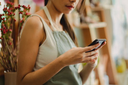 Free Crop anonymous florist in apron browsing mobile phone while working in cozy floral shop Stock Photo