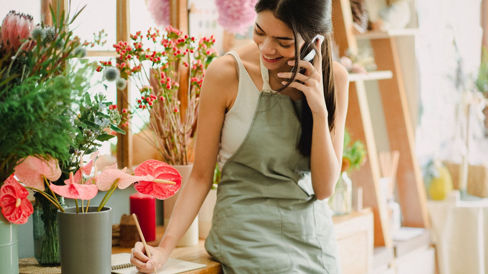 Smiling florist in a green apron takes a customer call on her smartphone and writes notes at the counter of her flower shop