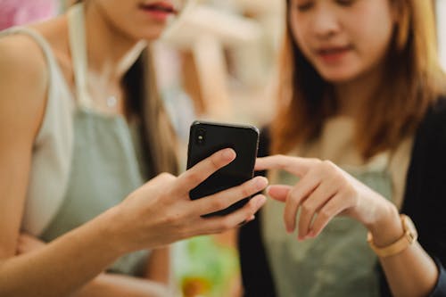 Crop faceless female florists in apron holding mobile phone while working in floral shop