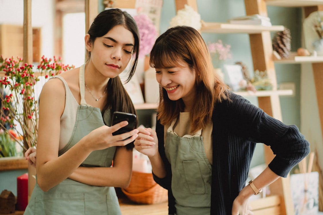 Free Cheerful multiracial female florists in aprons standing in flower shop and browsing mobile phone Stock Photo