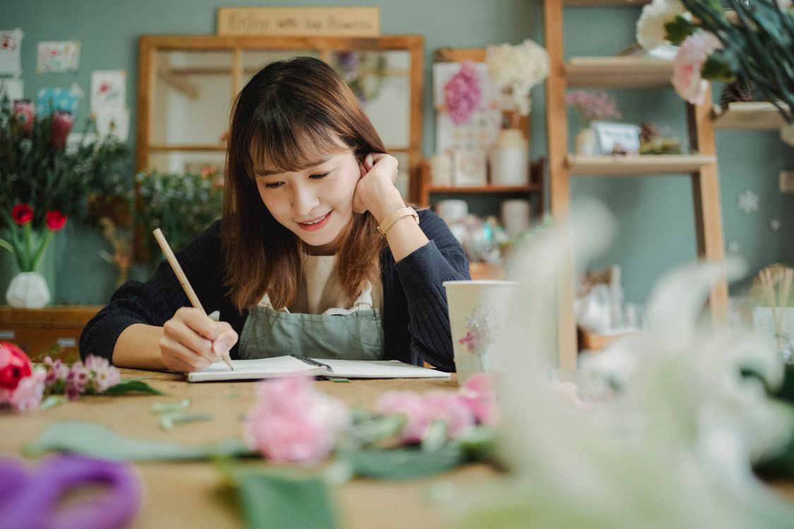 A florist jots notes in a notebook at a shop table covered with flowers, the kind of rough notes a good ChatGPT briefing starts from