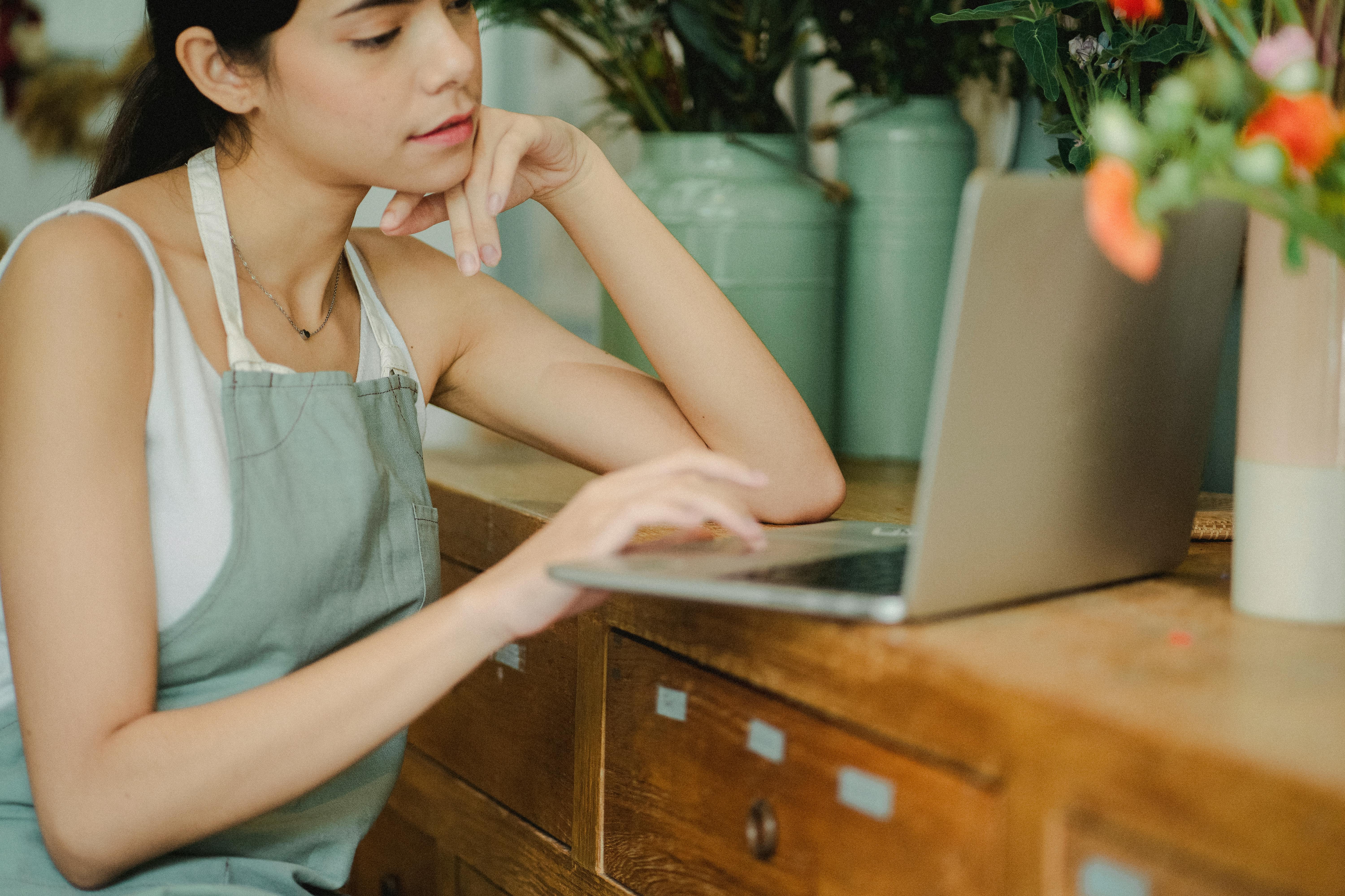 Crop female florist in apron sitting at table with laptop while working in cozy floral shop