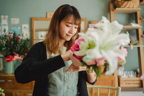 Smiling ethnic female florist in apron standing in floral shop and arranging bunch of fresh flowers