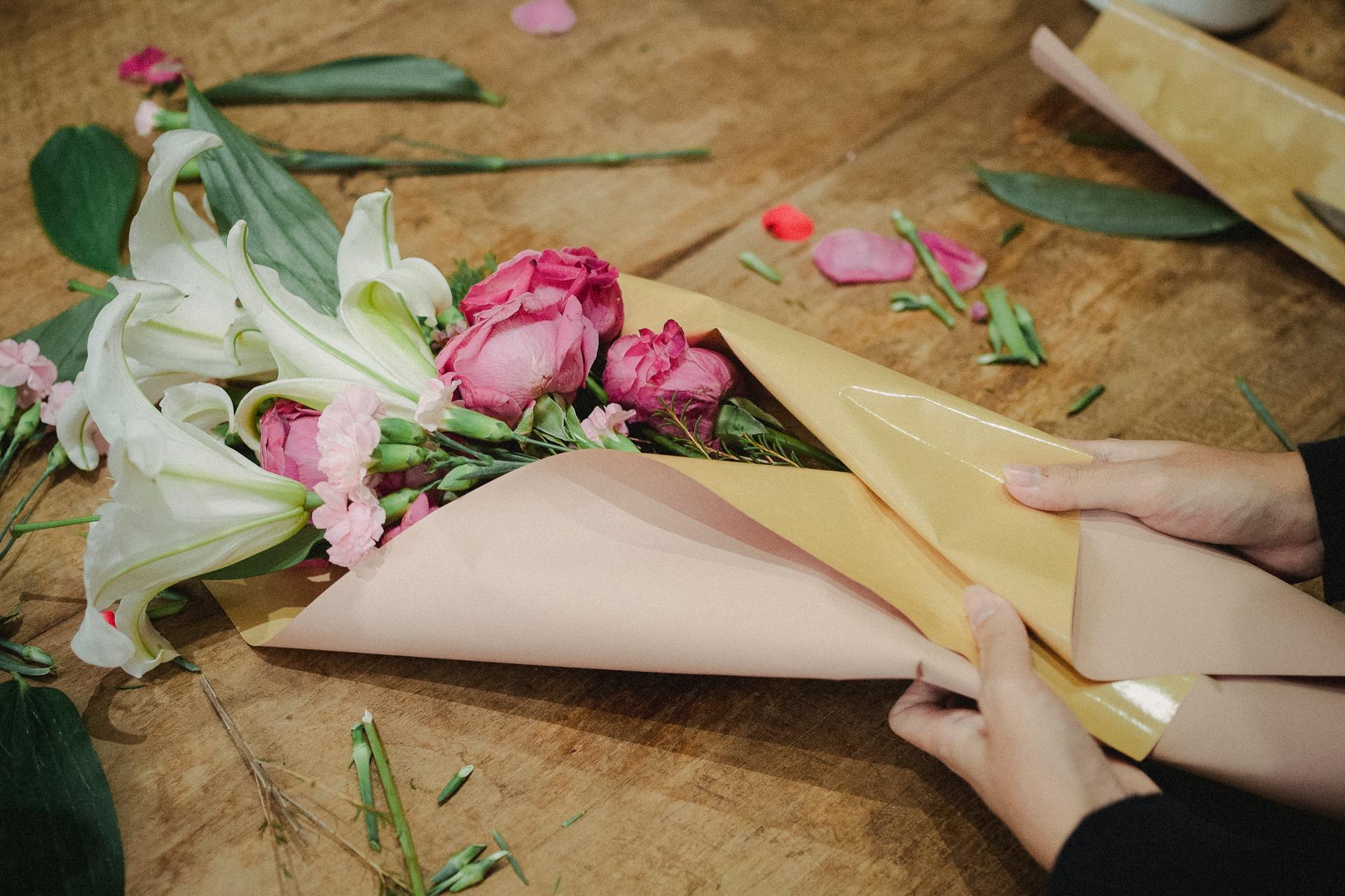 Hands wrapping a fresh floral bouquet with pink roses and white lilies on a wooden table.