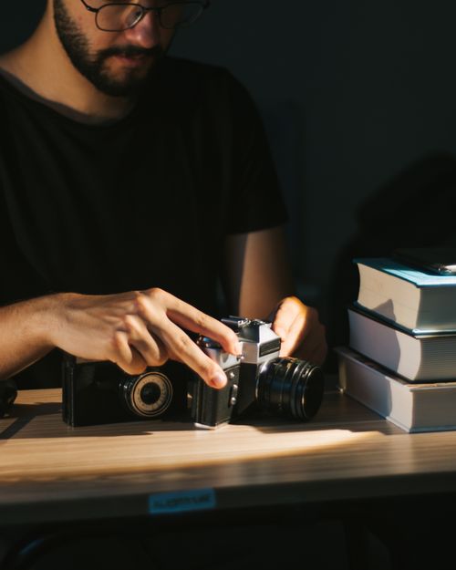 Free A man inspects a vintage camera on a desk next to a stack of books, highlighting creative exploration. Stock Photo