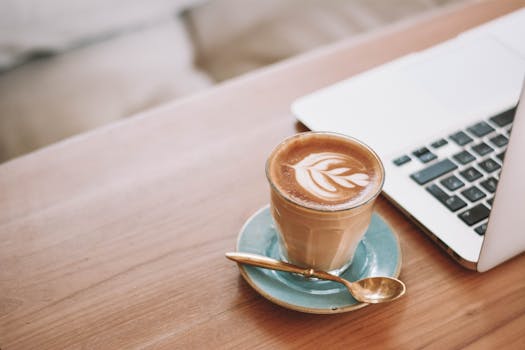 A glass of latte with intricate latte art next to a laptop on a wooden desk, perfect for digital nomad themes.