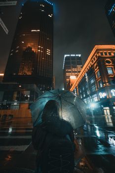 A person in silhouette with an umbrella under illuminated skyscrapers in Minneapolis's wet streets at night.