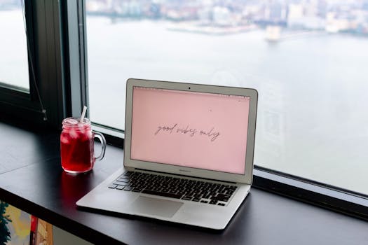 A laptop with 'good vibes only' text and a refreshing drink on a windowsill overlooking Jersey City.