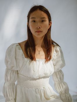 一位棕色头发、身着浪漫连衣裙的冷静年轻亚洲女士，在阳光下的白色背景前注视着镜头
