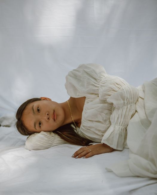 Pensive young ethnic woman in feminine dress lying on white bed sheet on sunny day and looking at camera