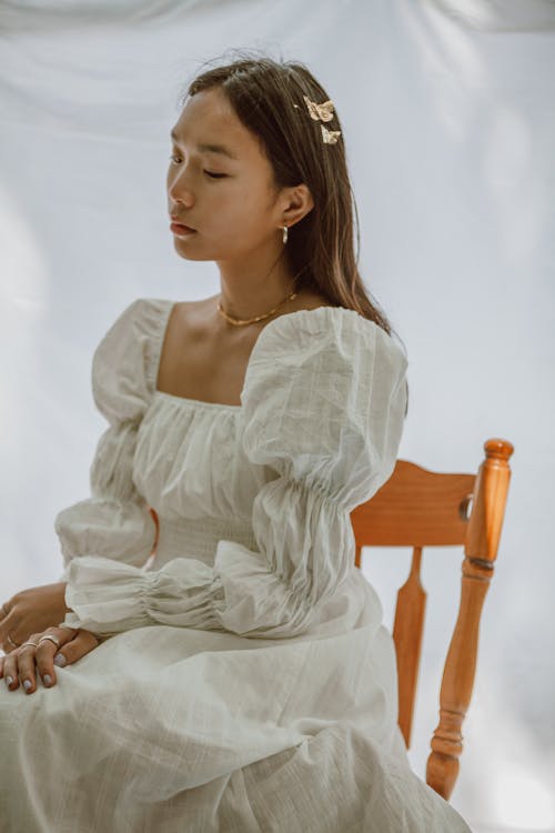 Side view of young contemplative Asian female in elegant white dress sitting on wooden chair and looking down thoughtfully
