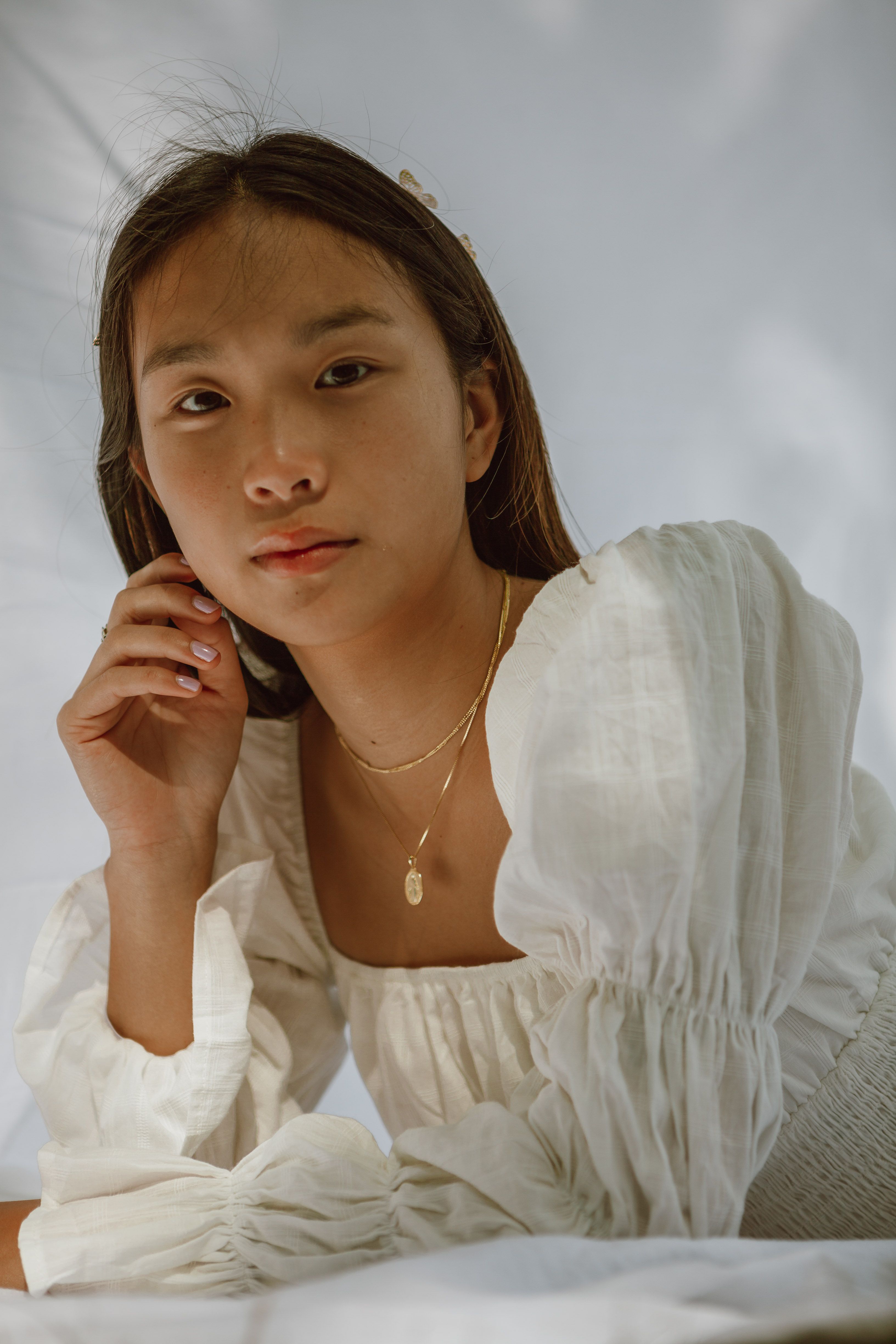 Elegant young Asian lady with perfect skin and long dark hair in stylish outfit touching face while lying on white blanket and looking at camera