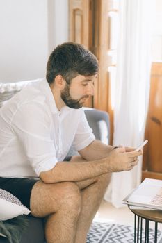 Adult man sitting on sofa using smartphone indoors with a relaxed posture.