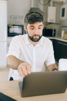 Man in dress shirt working on a laptop in a home kitchen setting, exemplifying remote work lifestyle.