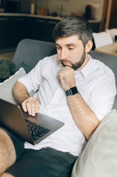 Caucasian man in deep thought, working on a laptop while sitting comfortably at home.
