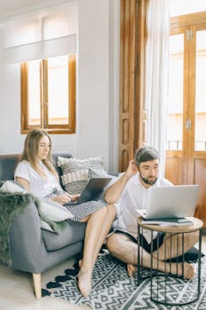 Caucasian couple working from home with laptops, sitting in a bright living room. Cozy and relaxed setting.