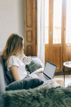 Woman holding coffee cup, using laptop on sofa, with natural indoor lighting.