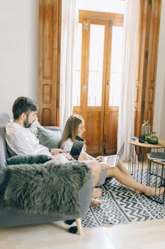 A couple engaging in remote work on laptops in a sunny living room setting.