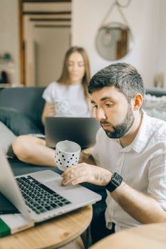 A couple works remotely with laptops in a cozy living room setting, enjoying coffee.