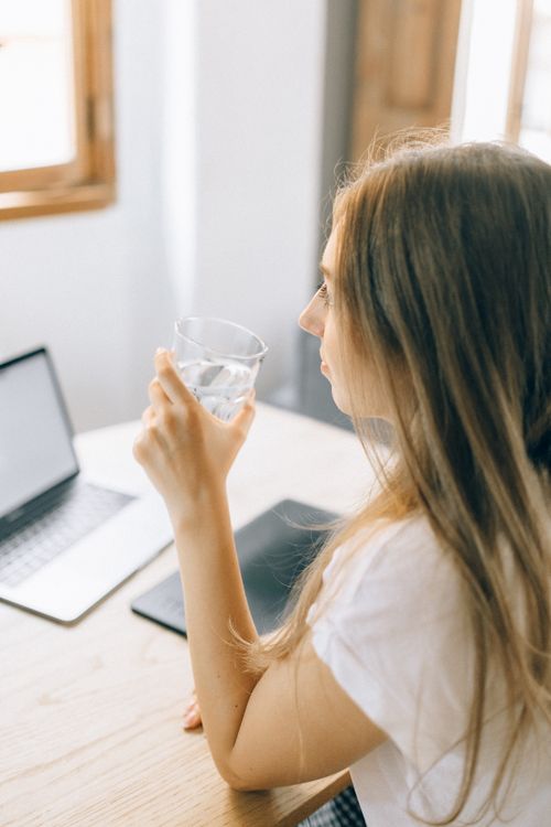 Free A woman in a white T-shirt holding a glass of water sits at a wooden table with a laptop, embodying relaxed home office vibes. Stock Photo