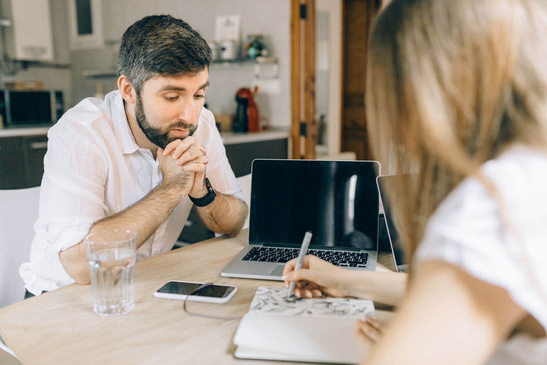 Casal jovem sentado à mesa da cozinha conversando com caderno e computador abertos.