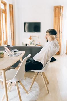 A man in a modern living room, relaxing with a laptop and TV, seated on a wooden chair.