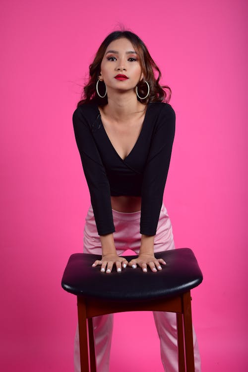 Free Dreamy ethnic lady with long hair and red lips wearing stylish clothes and looking at camera while standing on pink background with hands on chair Stock Photo