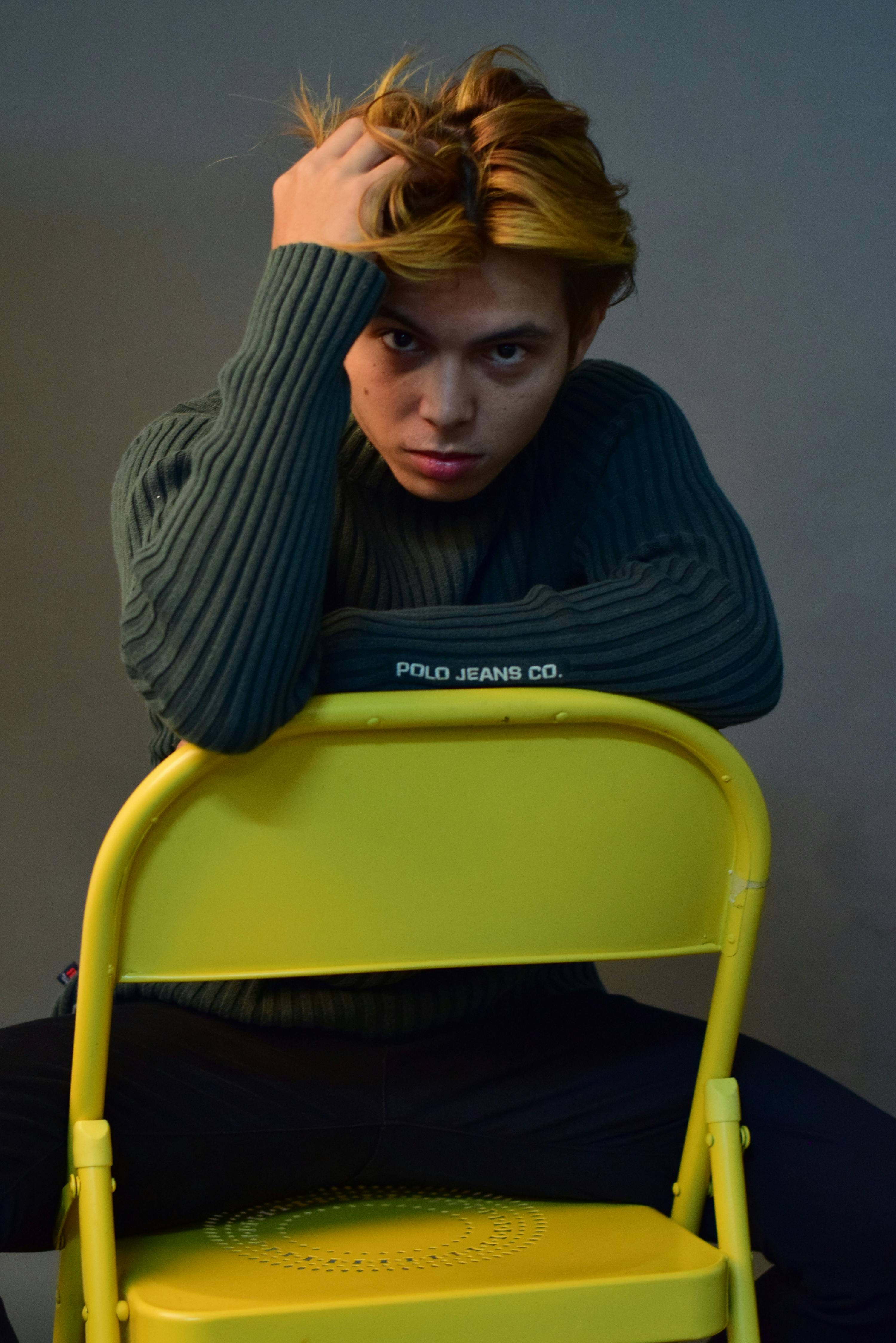 Thoughtful male with dyed hair wearing sweater while looking at camera and sitting on yellow chair in studio on gray background and touching hair