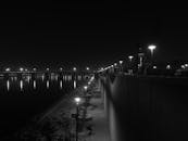 这张令人惊叹的黑白夜景照片中，一条光线昏暗的河畔小路上，明亮的城市灯光倒映在水面