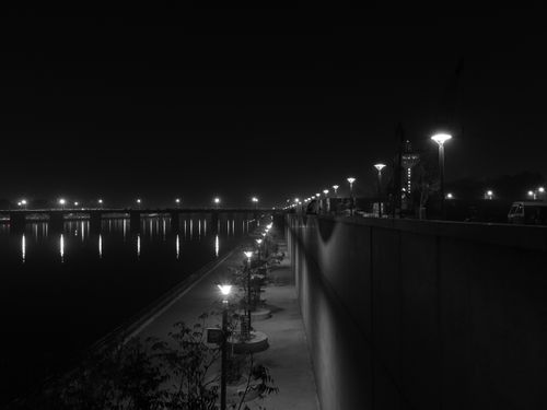 Free A stunning black and white night photo of a dimly lit riverside pathway with bright city lights reflecting on the water. Stock Photo