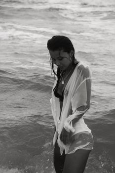 Black and white of young female with long dark hair in swimsuit and wet white shirt standing in water of ocean and looking down