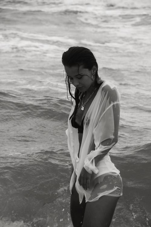 Free Black and white of young female with long dark hair in swimsuit and wet white shirt standing in water of ocean and looking down Stock Photo
