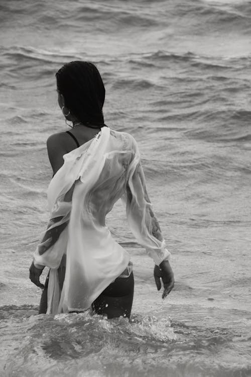 Black and white of anonymous female with dark hair in wet white shirt standing in waving sea