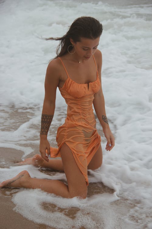 Young sensual female with brown hair and tattoos in wet tight dress kneeling on sandy coast of waving sea