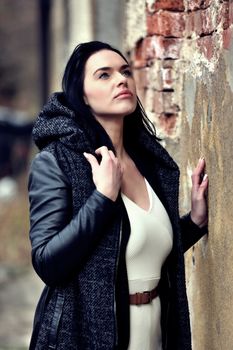 Elegant woman in black jacket and white dress looking up near a brick wall.