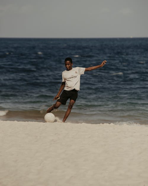 Young black man kicking ball on sandy beach