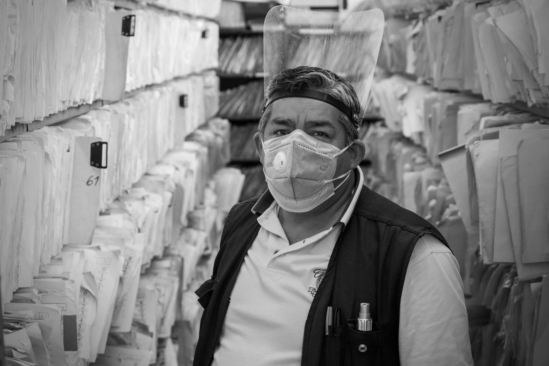 An elderly man with a face mask and shield in an archive aisle maintaining documents and records.