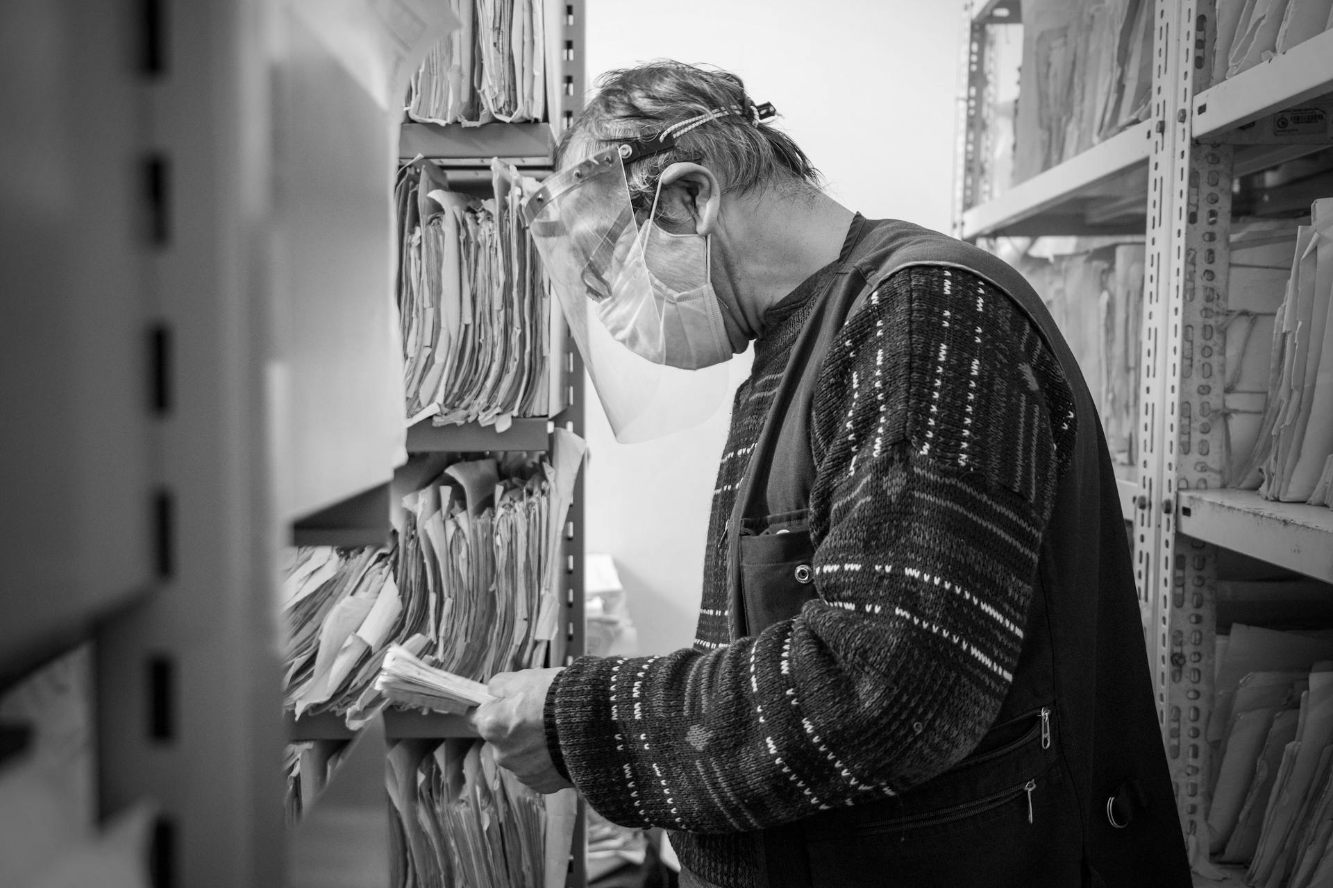 Elderly man wearing a face shield organizes files in a black and white office archive.