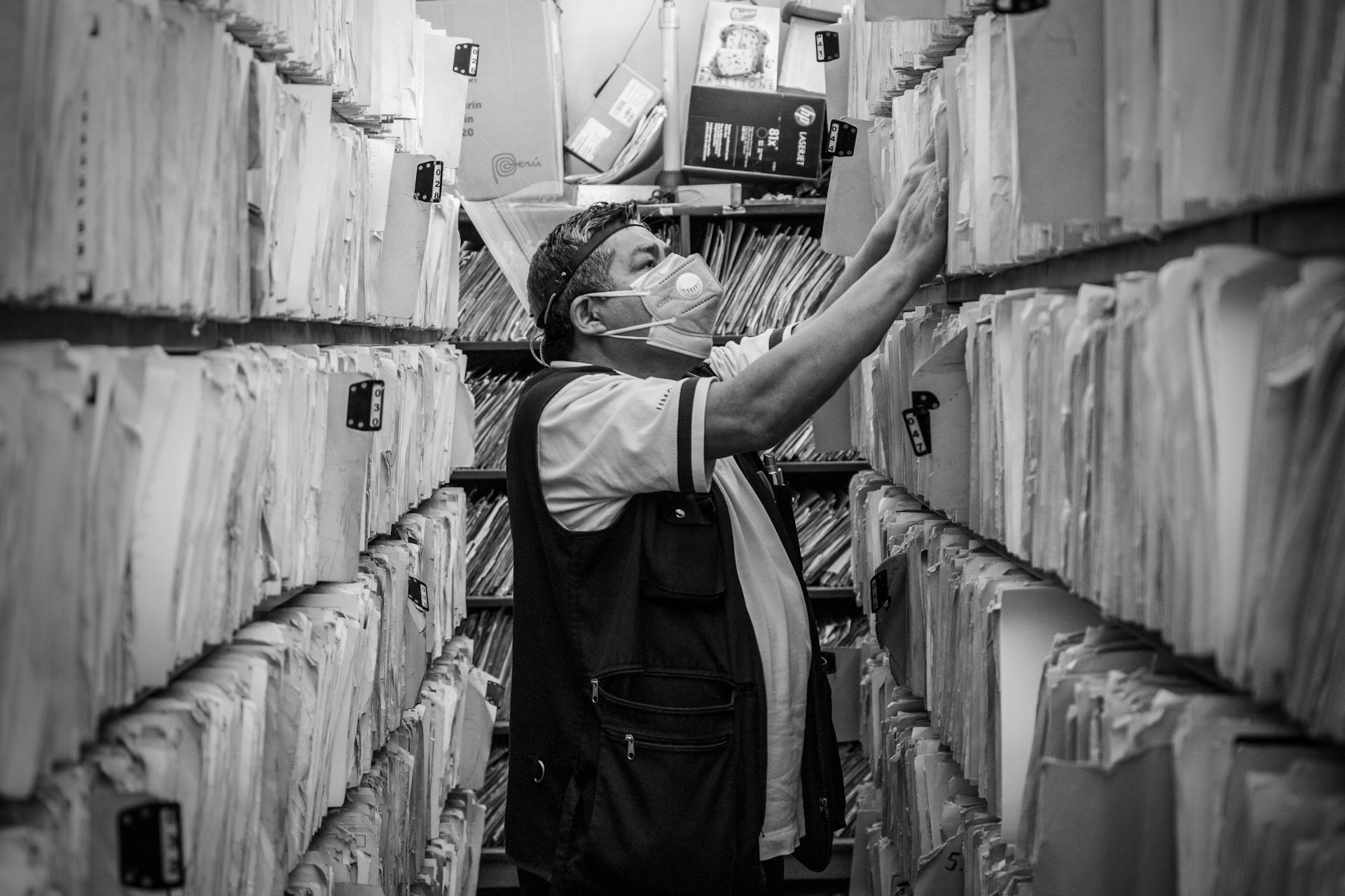 Elderly man sorts through files in an office aisle wearing a face mask.