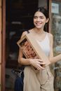 Cheerful lady in casual clothes with small cardboard package standing in doorway and looking away in city on daytime