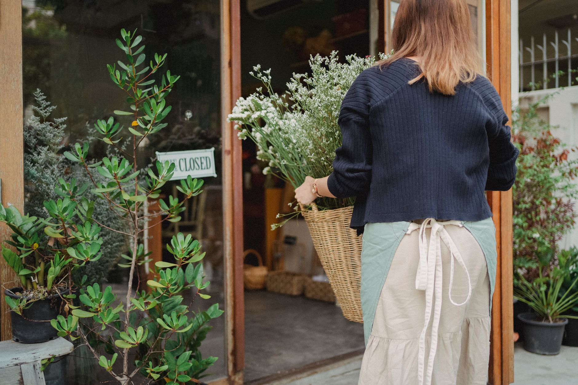 Back view of anonymous florist in apron carrying wicker basket of flowers to floral shop in daytime