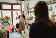阳光明媚的日子里，一位面带微笑的女人走进一家充满活力的花店，向店主挥手