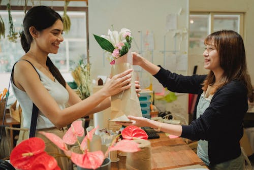 Side view of cheerful florist giving paper bag with bouquets of flowers to smiling client in floral shop